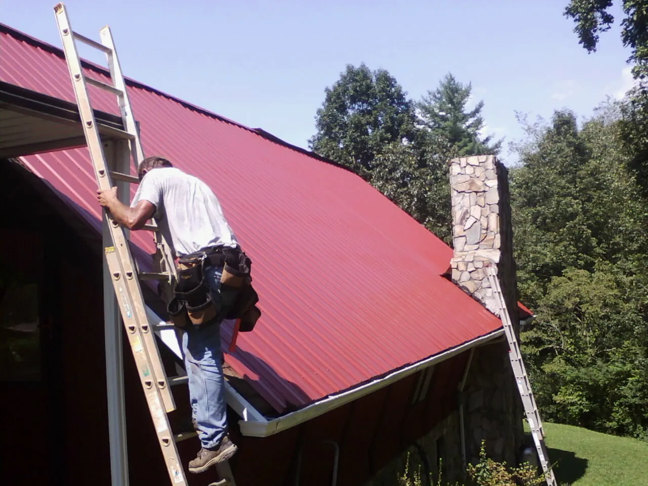 Roofer on a ladder beside a red metal roof and stone chimney — Soffit & Fascia Repair by Roofing Brothers in Annapolis